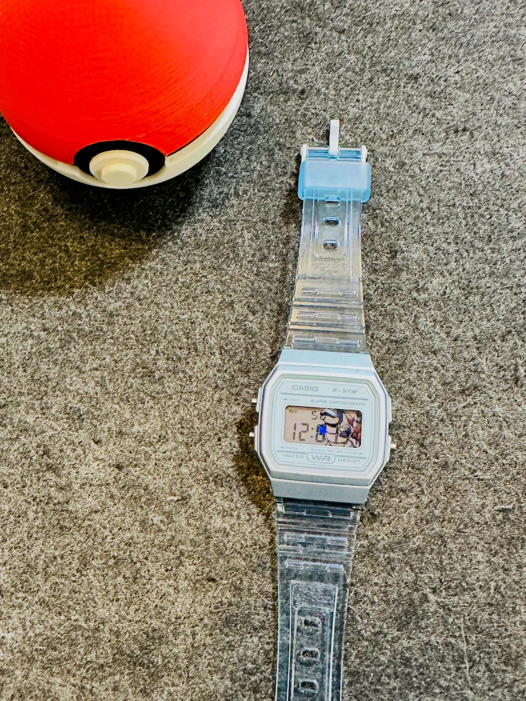 A Casio F-91W with a silver case and clear resin strap, a Blastoise print showing behind the LCD, beside an orange Poké Ball